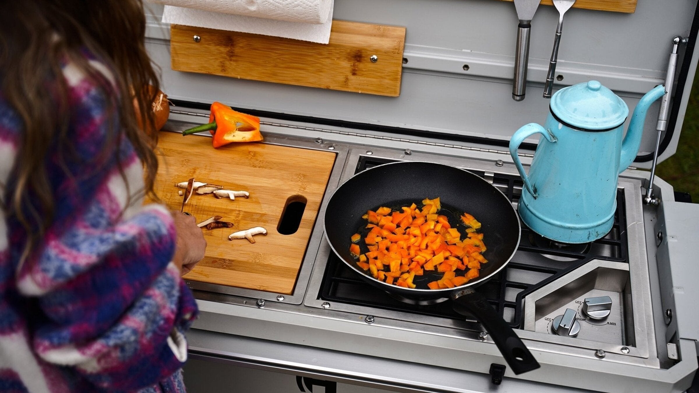 Camp Kitchen cooktop in use — frying pan with chopped carrots, utensils, and a teal kettle.